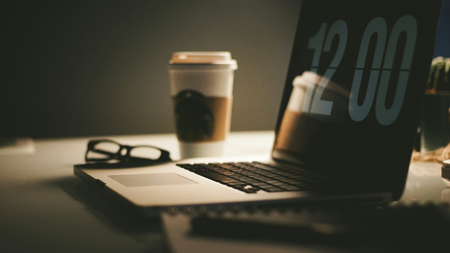 An open laptop, coffee cup, and reading glasses on a desk.